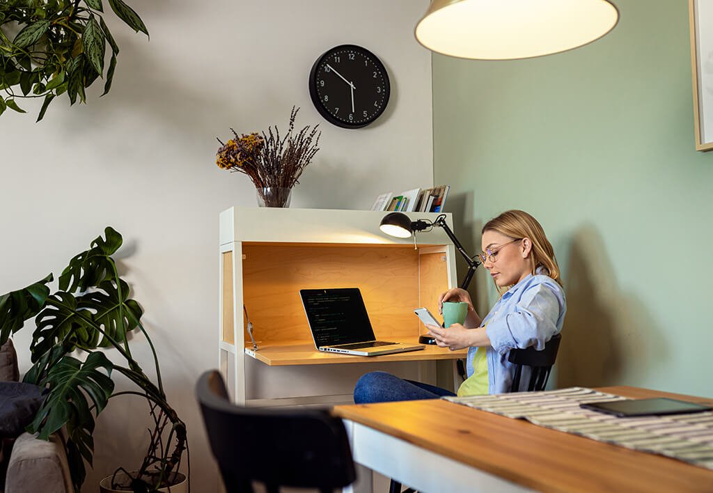 Shared Apartment Space | Woman Sitting at Fold Up Wall Desk Looking at Her Mobile Phone