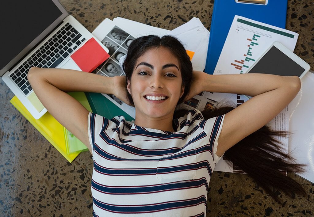 School and Work Balance | Woman Smiling While Lying On Her Back with Her Hand Behind Her Head, Lying On a Pile of School Work Papers